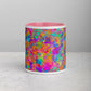 A front view of the Splotches and Crosses Mug by My Favourite Colour is Rainbow showing the pink rim and interior and featuring a bright, abstract design with splashes of neon pink, green, orange, and blue . The mug sits on a light grey surface on a plain background.