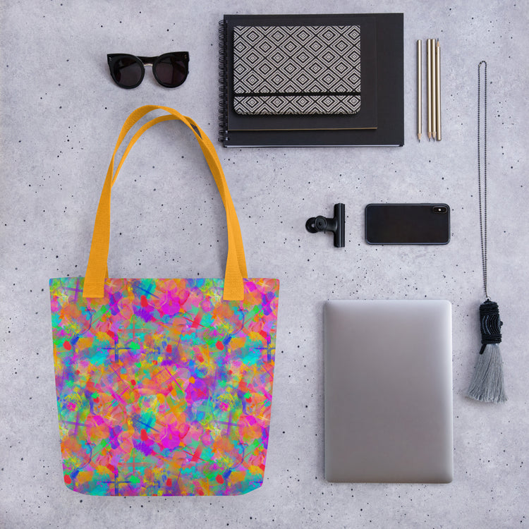 A flat lay of the vivid Splotches and Crosses Tote Bag by My Favourite Colour is Rainbow, featuring yellow handles, surrounded by sunglasses, a patterned notebook, pens, a phone, a camera attachment, a closed laptop, and a tassel accessory on a speckled surface.