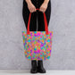 A person wearing black pants and boots carries a red-handled Splotches and Crosses Tote Bag by My Favourite Colour is Rainbow.  The bag features an abstract pattern of pink, blue, green, and orange splashes 