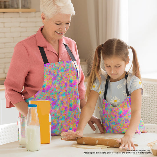 An elderly woman and a young girl, both wearing Splotches and Crosses Aprons by My Favourite Colour is Rainbow, bake together. The smiling woman watches as the girl rolls dough with a rolling pin. A bottle of milk and a cereal box are the table.  The apron features colourful abstract splashes of colour and bright cross shapes.