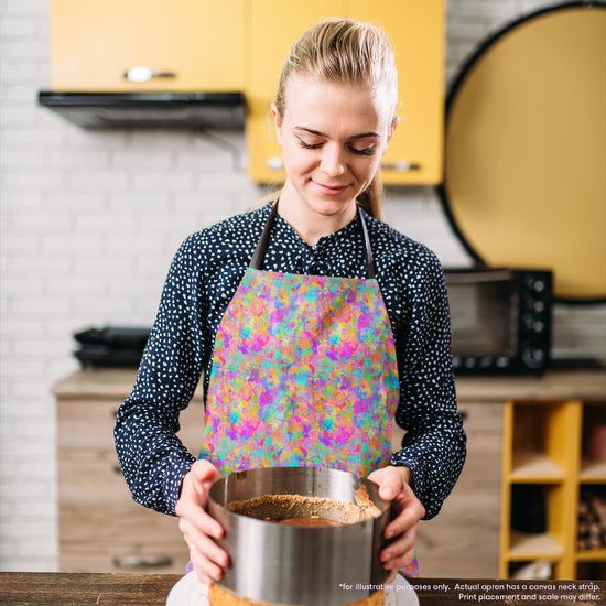 A woman wears a vibrant Splotches and Crosses Apron by My Favourite Colour is Rainbow over her polka dot blouse as she holds a round cake tin. Yellow cabinets and a microwave are in the background.  The apron features colourful abstract splashes of colour and bright cross shapes.