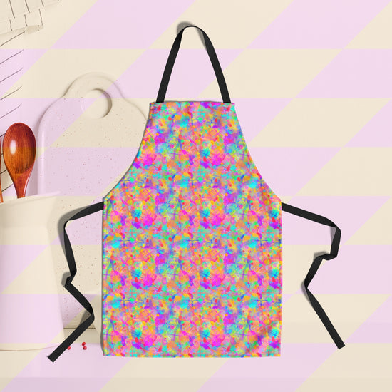 The Splotches and Crosses Apron by My Favourite Colour is Rainbow features a vibrant, abstract pattern of pink, aqua, and yellow splashes of colour. It has a black neck straps and ties. A white cutting board and wooden utensil holder are in the background.