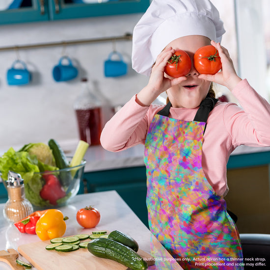 A child dons a chefs hat and a Splotches and Crosses Apron by My Favourite Colour is Rainbow, playfully holding two tomatoes over their eyes. The counter is full of veggies like cucumbers and bell peppers, complete with cooking tools and a wooden cutting board.  The apron features colourful abstract splashes of colour and bright cross shapes.