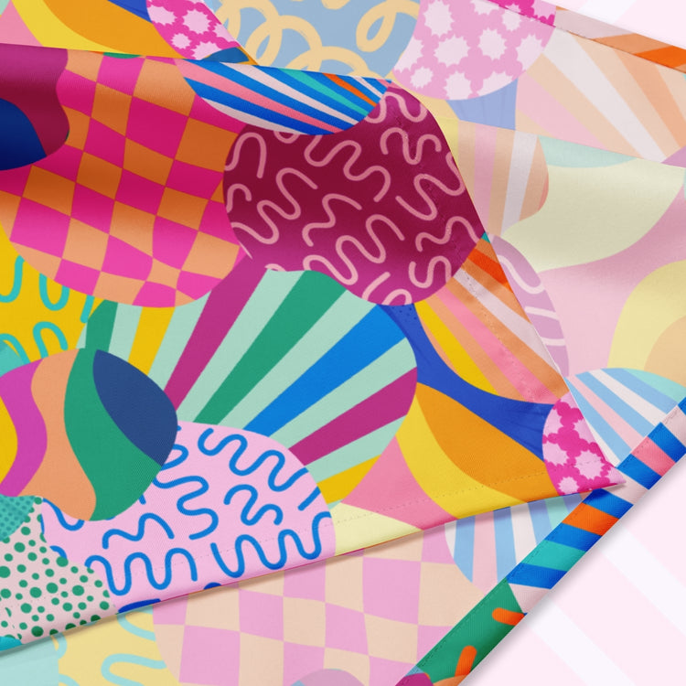 A closeup of the 'Squiggs and Stripes' Bandana by My Favourite Colour is Rainbow featuring a colourful abstract design with circles, waves, stripes, and checkered patterns in pink, blue, green, yellow, and orange. The design is printed on only one side of the bandana.