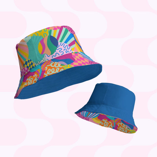 The Squiggs and Stripes Reversible Bucket Hat by My Favourite Colour is Rainbow features bold abstract patterns in pink, green, yellow, and blue on one side, and a solid blue design with a patterned rim. Displayed against a light background with pink and white geometric shapes, its perfect for matching your Style BFF in multiple sizes.