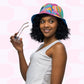 A woman with curly hair smiles, holding eyeglasses and wearing the Squiggs and Stripes Reversible Bucket Hat by My Favourite Colour is Rainbow with a white sleeveless top. The light pink and white geometric background enhances her as the ultimate Style BFF.