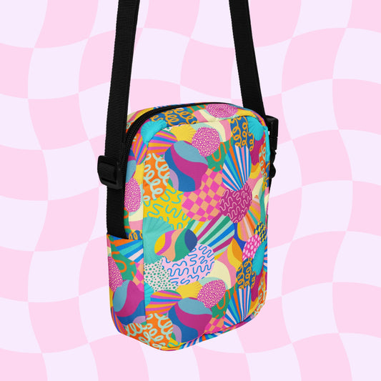 The Squiggs and Stripes Mini Crossbody bag by My Favourite Colour is Rainbow features vibrant swirls, patterns, and shapes in pink, green, yellow, and blue tones. It has sturdy adjustable black straps set against a pink and white checkered background.