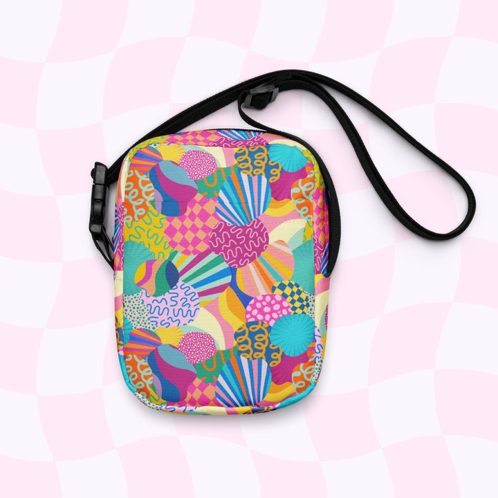 The Squiggs and Stripes Mini Crossbody Bag by My Favourite Colour is Rainbow is a small, water-resistant crossbody with colorful abstract swirls, stripes, and circles in bright hues. It features sturdy black adjustable straps on a pink and white checkered background.