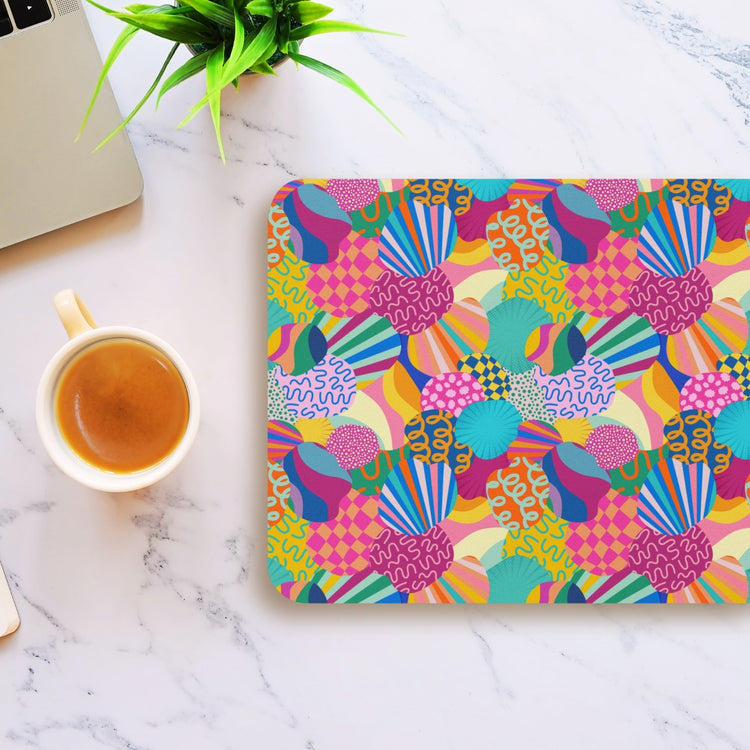 The Squiggs and Stripes Mouse Pad by My Favourite Colour is Rainbow displays vibrant abstract patterns on a marble desk. A laptop peeks in from the left, while a coffee cup rests near the pad, accompanied by a small green plant in the corner.