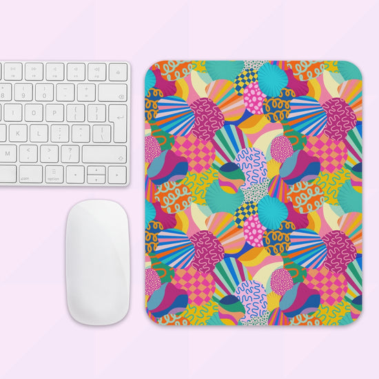 The light pink background showcases the Squiggs and Stripes Mouse Pad by My Favourite Colour is Rainbow, featuring an abstract pattern in blue, pink, yellow, and green alongside a white keyboard and mouse.