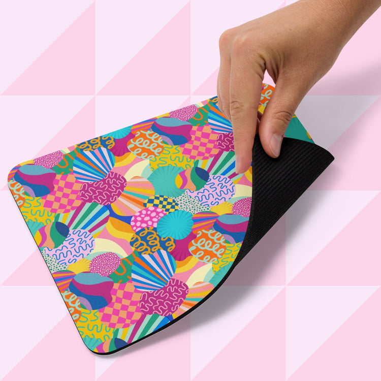 A hand displays the Squiggs and Stripes Mouse Pad by My Favourite Colour is Rainbow, featuring a vibrant design of abstract, geometric shapes in pink, yellow, green, and blue hues on a pink and white triangular background.