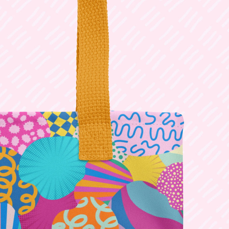 The Squiggs and Stripes Tote Bag by My Favourite Colour is Rainbow features a mustard yellow handle, set against a vibrant abstract pattern of blue, pink, orange, green, and purple swirls and circles. The design pops on a light pink background.