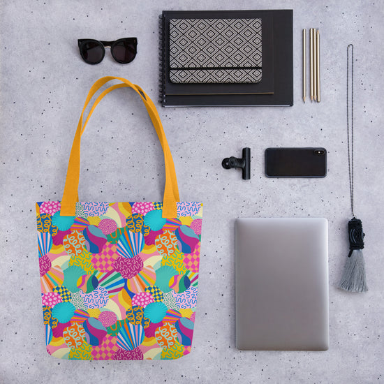 A flat lay on a gray speckled surface showcases the Squiggs and Stripes Tote Bag by My Favourite Colour is Rainbow, sunglasses, two notebooks, a patterned pouch, three pens, a phone, a clip, a tassel keychain, and an on-demand laptop.