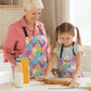 In a bright kitchen, a woman and young girl enjoy cooking together. They wear vibrant Squiggs and Stripes Aprons from My Favourite Colour is Rainbow as the girl rolls dough, her grandmother smiling. A milk bottle and baking supplies are spread across the table. The apron features abstract colourful shapes, including checks, stripes, squiggles and more, in rainbow colours