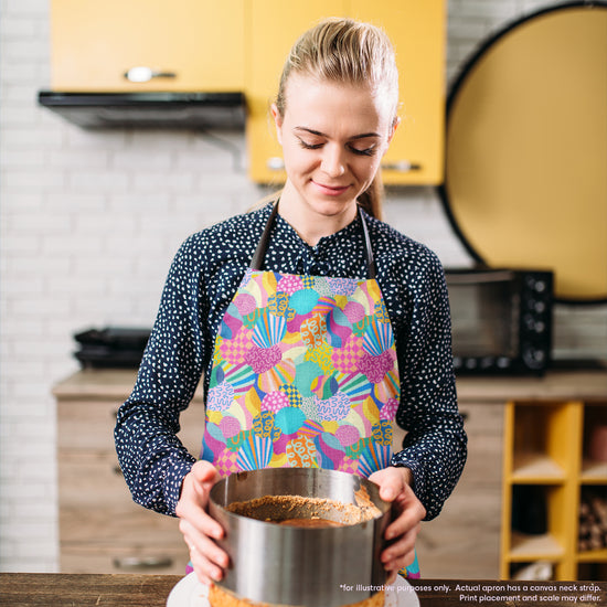 In a kitchen with yellow cabinets, a person wearing the Squiggs and Stripes Apron by My Favourite Colour is Rainbow holds a baking pan with crust, looking down at their work. The kitchen includes a microwave and other essentials, showcasing their cooking experience.  The apron features abstract colourful shapes, including checks, stripes, squiggles and more, in rainbow colours