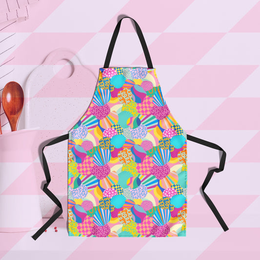 The vibrant Squiggs and Stripes Apron by My Favourite Colour is Rainbow features abstract colourful shapes, including checks, stripes, squiggles and more, in rainbow colours.  It has a black strap and ties. A white cutting board and wooden spoons are in the background.