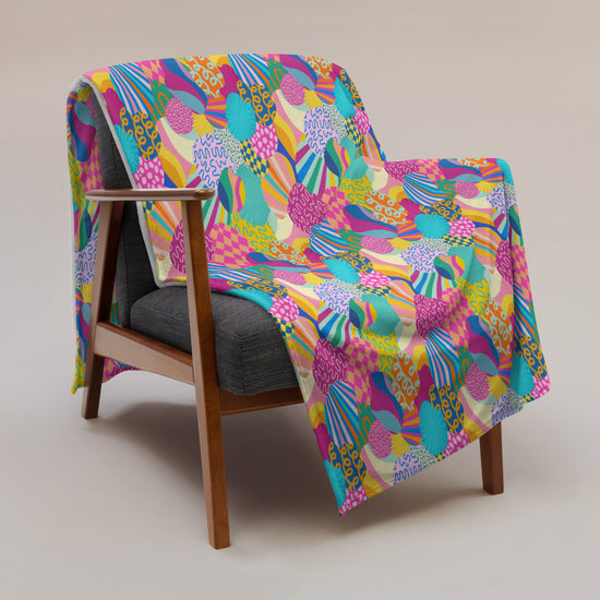 Featuring vibrant abstract patterns in pink, blue, yellow, and green, the Squiggs and Stripes Throw Blanket by My Favourite Colour is Rainbow adds a cosy touch to a mid-century modern wooden chair with a gray cushion.