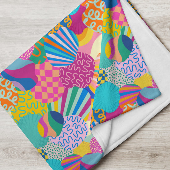 The Squiggs and Stripes Throw Blanket by My Favourite Colour is Rainbow rests on a wooden surface, featuring abstract spirals, shells, circles, and zigzags in vibrant pink, blue, yellow, and green.