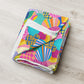 The Squiggs and Stripes Throw Blanket by My Favourite Colour is Rainbow, featuring a vibrant abstract design with stripes and swirls in pink, yellow, blue, and green, rests on a light wood surface.