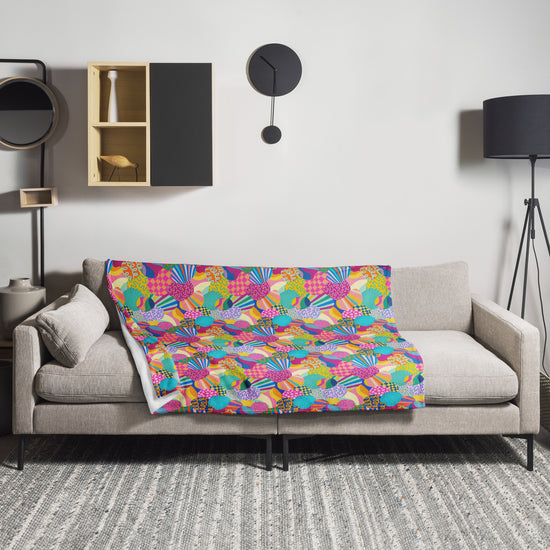 A beige sofa with a Squiggs and Stripes Throw Blanket by My Favourite Colour is Rainbow enhances the rooms design. A modern clock and square shelving adorn the wall, with decorative items, while a tall floor lamp stands to the right, and a gray patterned rug creates a cozy atmosphere.