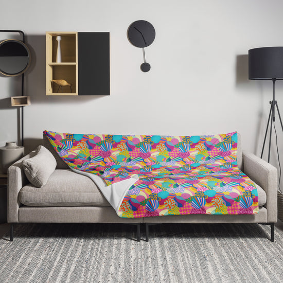 A modern living room showcases a gray sofa with the Squiggs and Stripes Throw Blanket from My Favourite Colour is Rainbow, boasting vibrant designs. A black floor lamp, round wall clock, and wall shelf enhance the cozy ambiance, while a gray rug seamlessly ties the space together.