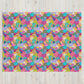 The Squiggs and Stripes Throw Blanket by My Favourite Colour is Rainbow features a vibrant design of overlapping geometric shapes, stripes, and polka dots in pink, blue, yellow, and green on a light wooden background.