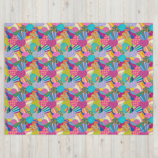 The Squiggs and Stripes Throw Blanket by My Favourite Colour is Rainbow features a vibrant design of overlapping geometric shapes, stripes, and polka dots in pink, blue, yellow, and green on a light wooden background.