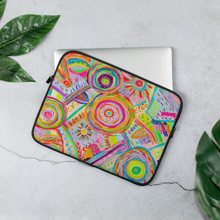 The 13 inch Summer Sorbet Laptop Sleeve by My Favourite Colour is Rainbow partially covers a grey laptop on a light surface. The colourful features abstract patterns with bright circles. Green leaves are artfully arranged around it.