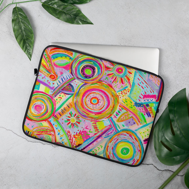 The 15 inch Summer Sorbet Laptop Sleeve by My Favourite Colour is Rainbow partially covers a grey laptop on a light surface. The colourful features abstract patterns with bright circles. Green leaves are artfully arranged around it.