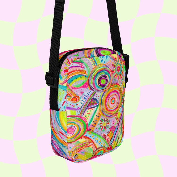 The Summer Sorbet Mini Crossbody Bag by My Favourite Colour is Rainbow boasts a vibrant abstract pattern with swirling pink, green, orange, and blue designs. It features adjustable black straps against a pastel pink and light green checkered backdrop.