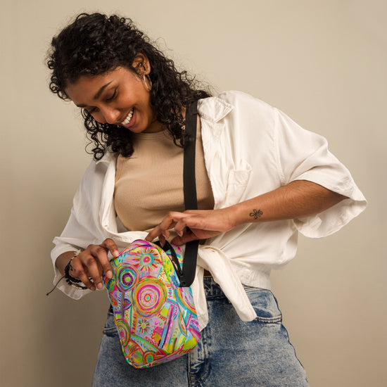 A woman with curly hair smiles, dressed in a white shirt over a tan top and denim jeans. Against a neutral backdrop, she adjusts her My Favourite Colour is Rainbow Summer Sorbet Mini Crossbody Bag with adjustable straps. A tattoo graces her forearm.