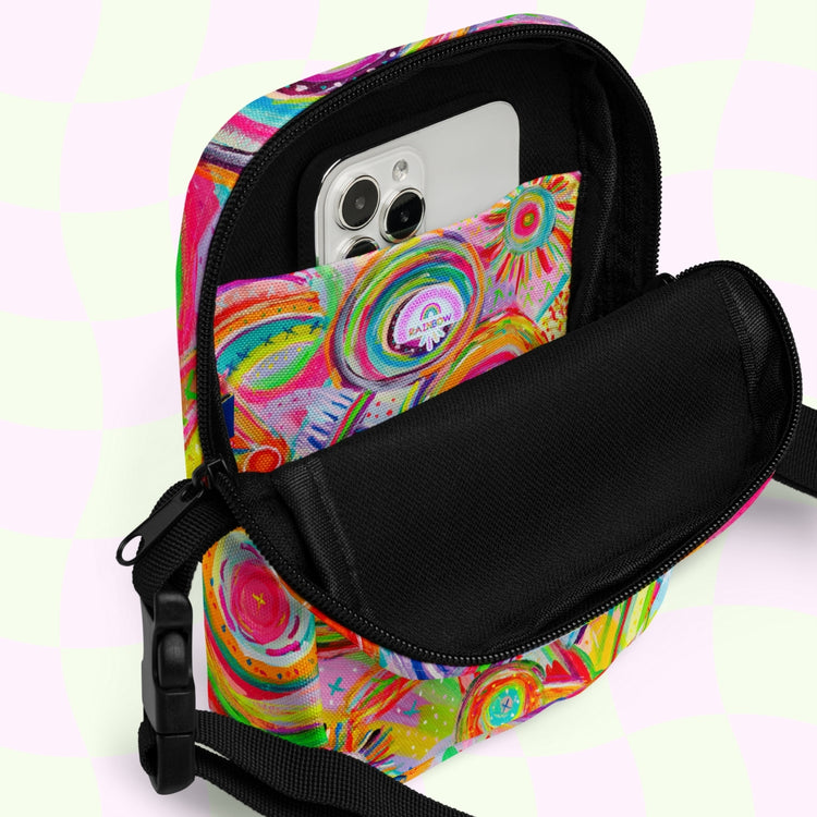 The My Favourite Colour is Rainbows Summer Sorbet Mini Crossbody Bag showcases a vibrant abstract print and open design with a smartphone in the front pocket, set against a light checkered background. It features adjustable straps for on-the-go comfort.