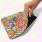 A hand lifts the Summer Sorbet Mouse Pad by My Favourite Colour is Rainbow, revealing its black underside. The vibrant design showcases circles, abstract shapes, and bright colors like pink, green, and blue on a pale patterned background.