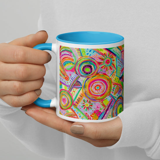 A person holds the Summer Sorbet Mug by My Favourite Colour is Rainbow, showcasing a vibrant abstract design with colorful circles and patterns. The mug features a bright blue interior and handle.