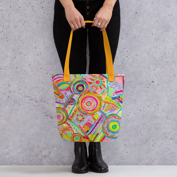 Someone holds the trendy Summer Sorbet Tote Bag by My Favourite Colour is Rainbow, showcasing a vibrant abstract circle and line pattern with yellow handles. They're styled in black pants and boots, standing against a gray speckled background.