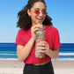 A woman in a red shirt and sunglasses joyfully holds her colorful Summer Sorbet Stainless Steel Tumbler by My Favourite Colour is Rainbow, complete with a straw. She stands on a sunny beach with the ocean glistening under the clear blue sky.