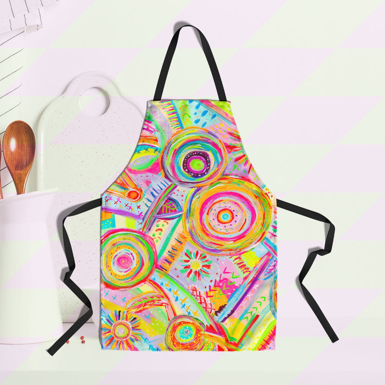 The Summer Sorbet Apron by My Favourite Colour is Rainbow features abstract colourful circles, lines, and geometric patterns in neon hues. Displayed against a white kitchen background, it inspires culinary creativity with a cutting board and utensils visible nearby.