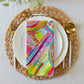 A vibrant napkin from the Mixed Set of 4 Art Napkins by My Favourite Colour is Rainbow sits on a white plate, resting atop a woven placemat. Gold cutlery flanks the plate, offering a chic dining upgrade on a light-textured surface.