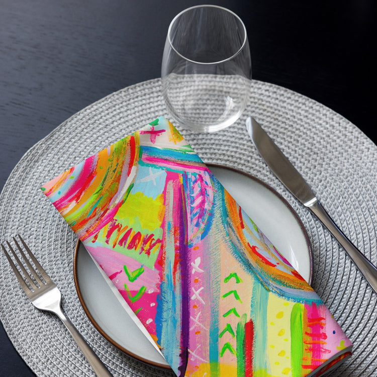 A Summer Sorbet-patterned napkin from My Favourite Colour is Rainbow on a white plate. A knife and fork sit on a grey woven placemat, while a clear wine glass stands above the plate.