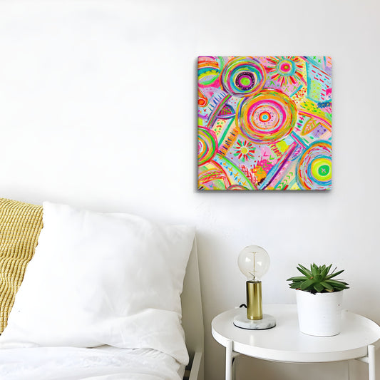 A vibrant 12x12 canvas artwork by My Favourite Colour is Rainbow is mounted on a white wall. Below, a white bed with a yellow pillow sits next to a small round table with a lightbulb lamp and potted plant.