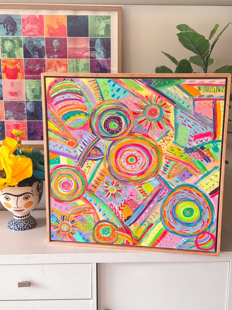 The Summer Sorbet Framed Original Painting by My Favourite Colour is Rainbow features vibrant circular patterns and lines, complemented by colorful portraits in a square frame and a whimsical ceramic face vase with yellow flowers. A green plant adds to the charm of this acrylic on canvas piece.