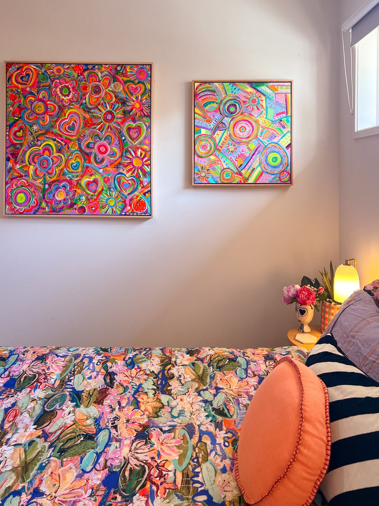 A colorful bedroom boasts the Summer Sorbet framed original painting by My Favourite Colour is Rainbow, a floral-patterned bedspread, and an orange round pillow. A small table with a glowing lamp and pink flowers sits in the corner, as natural light filters through the window.