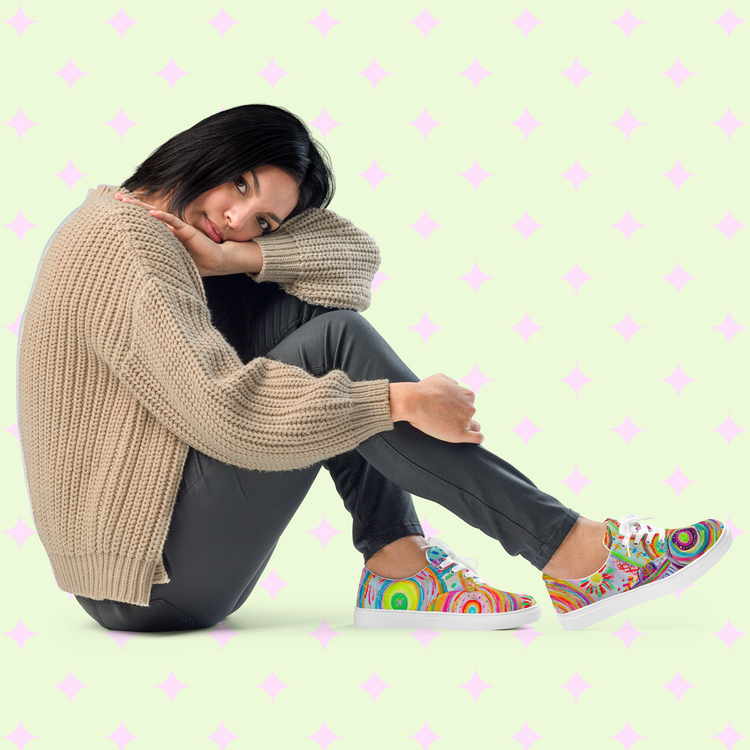 A person with short dark hair sits on the floor, resting their head on their knee. Theyre wearing a beige sweater, gray pants, and vibrant Summer Sorbet Canvas Sneakers by My Favourite Colour is Rainbow. The light green background with pink star patterns adds a playful vibe.