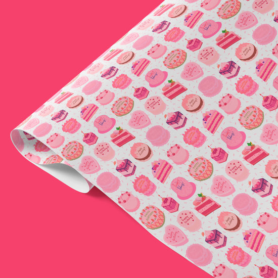 Sweet Treats Wrapping Paper Sheets by My Favourite Colour is Rainbow features  illustrations of decorated birthday cakes and desserts in shades of pink with green accents. The sheet is partially rolled out.