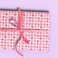 A gift is wrapped in Sweet Treats Wrapping Paper by My Favourite Colour is Rainbow, featuring illustrations of decorated birthday cakes and desserts in shades of pink with green accents.  It has a pink bow.