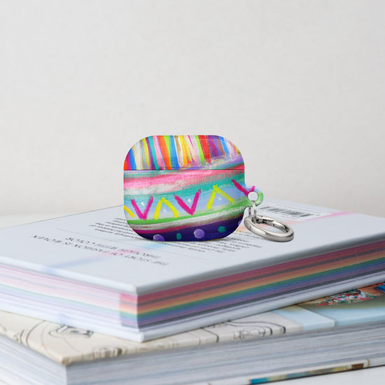 The Rainbow Connection Airpods Gen 3 Case stands on a stack of books.  It features  an abstract design of colourful green, pink, yellow, blue, and purple stripes, dots, and zigzags. 
