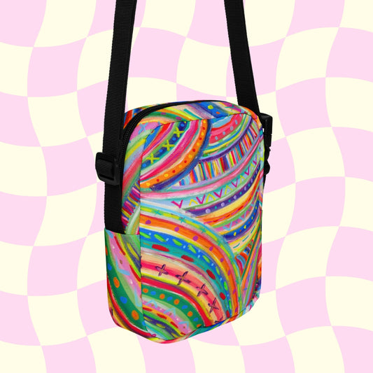 The Rainbow Connection Mini Crossbody Bag by My Favourite Colour is Rainbow features a vibrant psychedelic pattern with swirls and dots in pink, orange, green, and blue. Its displayed on a pink and cream checkerboard background with black adjustable straps and side accents.