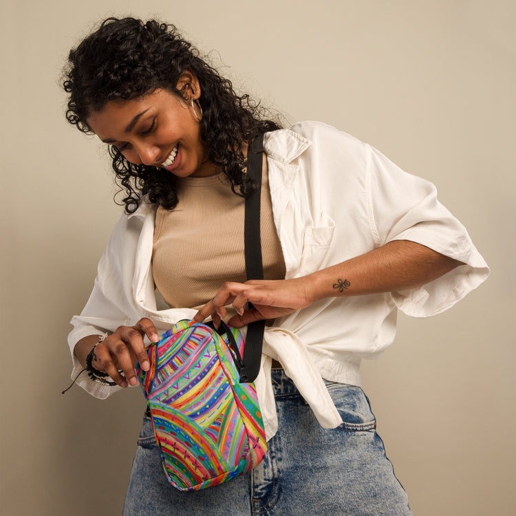 A person with curly hair smiles as they open The Rainbow Connection Mini Crossbody Bag by My Favourite Colour is Rainbow. With adjustable straps, it complements their beige top, white open shirt, and blue jeans against a plain background.