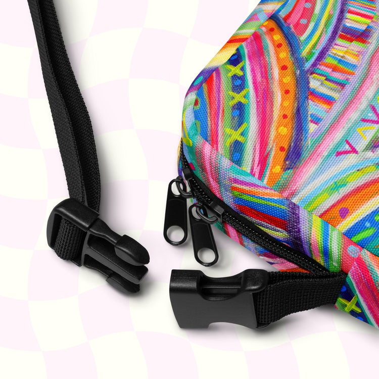 Close-up of The Rainbow Connection Mini Crossbody Bag by My Favourite Colour is Rainbow, showcasing intricate patterns in pink, blue, yellow, and green. The bag features a black zipper with dual sliders, adjustable straps, and black buckles against a light checkered background.