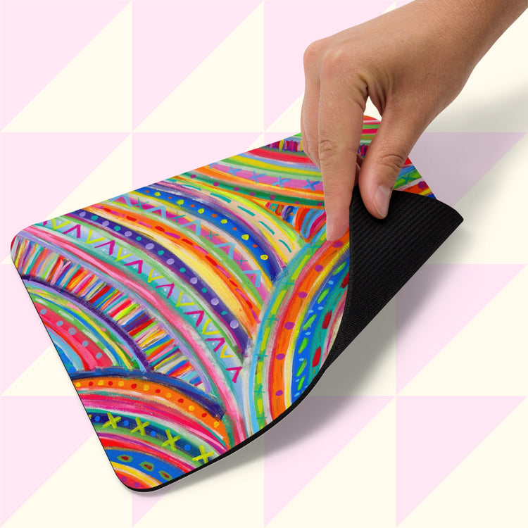 A hand lifts the corner of The Rainbow Connection Mouse Pad by My Favourite Colour is Rainbow, set against a soft pink geometric background. The mouse pad features vibrant multicolored stripes and dots, creating a lively and playful design.